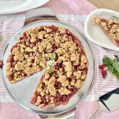 Ein köstlicher Rhabarberkuchen mit Streuseln, garniert mit kleinen Gänseblümchen. Frischer Rhabarber liegt daneben.