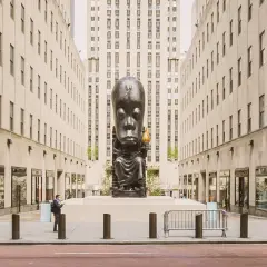 A monumental sculpture of a seated figure with a large head, holding a torch, stands as a striking example of public art between two imposing buildings.