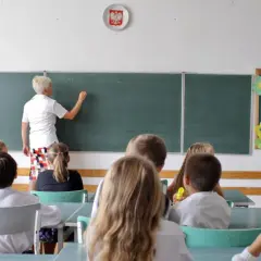 Nauczycielka pisze na tablicy, a uczniowie siedzą w ławkach. To scena z lekcji, która mogłaby się odbywać tuż przed tym, jak skończyło się gimnazjum w Polsce.