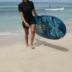 Kobieta w czarnej piance do pływania damskiej trzyma deskę surfingową z motywem liści palmowych na piaszczystej plaży.