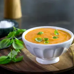 En skål med krämig rostad tomatsoppa, garnerad med färsk basilika och en skvätt olivolja. Tillbehör som salt och pepparkvarn syns i bakgrunden.