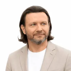 Mężczyzna z siwiejącą brodą i długimi włosami, ubrany w jasną marynarkę, patrzy w bok. Jego **majdan wzrost** jest imponujący.