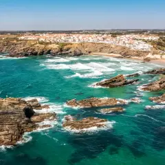 Costa rocosa y aguas turquesas en una **playa Portugal**. Un pueblo blanco se asoma al acantilado.