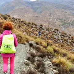 Senderista con mochila amarilla en un sendero montañoso. El grupo avanza, algunos a caballo, en un paisaje árido.