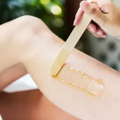 En hand applicerar vax med en träspatel på ett ben för att vaxa bikinilinjen hemma.