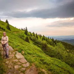 Kobieta z kijkami na kamiennym szlaku, podziwia widok na zielone zbocze i las. Widać, ile się wchodzi na Śnieżkę.