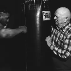 Mike Tyson trains with his manager, a bald man in a plaid shirt, in a dimly lit gym.