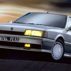 Un elegante Renault 21 Turbo plateado, con sus faros amarillos encendidos, se luce contra un cielo crepuscular.