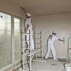 Dwóch fachowców w białych kombinezonach, używając kielni, wyrównuje ściany gładzią.