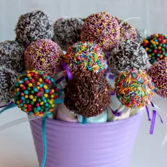 Kolorowe cake pops z jasnego biszkoptu, udekorowane posypkami i wiórkami kokosowymi, w fioletowym wazonie.