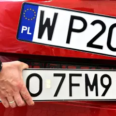 Ręka z bransoletką montuje tablicę rejestracyjną z napisem "O 7FM91" na czerwonym samochodzie. Obok widoczna jest inna tablica "W P20".