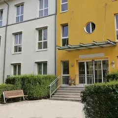 Gelbes Gebäude mit runden Fenster, Eingang mit Glasdach und Stufen. Ein Ort, der §43b SGB XI-Leistungen bietet, lädt zum Verweilen ein.