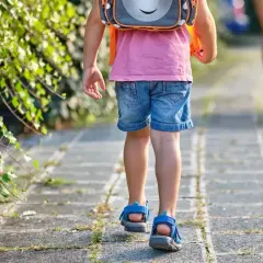 Małe dziecko w różowej koszulce i dżinsowych szortach z plecakiem idzie chodnikiem. To widok, kiedy dzieci idą do szkoły.