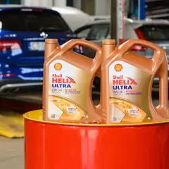Dwa kanistry oleju silnikowego Shell Helix Ultra 0W-30 stoją na czerwonym bębnie. W tle widać samochody w warsztacie.