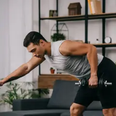 Mężczyzna wykonuje trening oporowy w domu, podnosząc hantle, opierając się o krzesło.