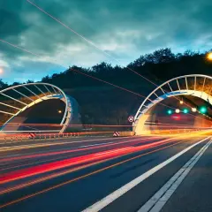 Dwa tunele drogowe oświetlone nocą, z rozmazanymi światłami samochodów tworzącymi smugi. Wjazd do tunelu po prawej stronie ma znak ograniczenia prędkości 60. Dawna opłata drogowa już nie obowiązuje.