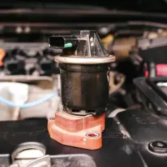 Componente negro con conector eléctrico, posiblemente una válvula de admisión, sobre una base rojiza en un motor.