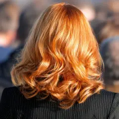 Kobieta z rudymi, falowanymi włosami w czarnej marynarce, na tle rozmytego tłumu.