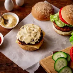 Pyszne hamburgery w trakcie przygotowania. Na stole sosy, bułki i świeże ogórki do hamburgerów.