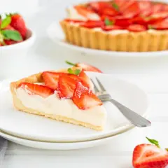 Pyszna tarta truskawkowa z kremowym nadzieniem i świeżymi owocami. Kawałek ciasta na talerzyku, obok cała tarta i miseczka truskawek.