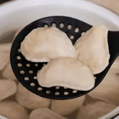Gotowane pierogi na czarnej łyżce cedzakowej w garnku z wodą.
