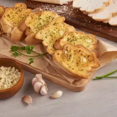 Chrupiące grzanki z piekarnika, posypane ziołami i czosnkiem, obok miseczki z posiekanym czosnkiem i masłem czosnkowym.