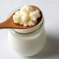 En träsked med vita kefir-korn vilar ovanpå en glasburk fylld med mjölk. Kefir nyttigt för magen!