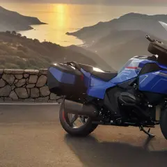 Una moto blu pronta per un viaggio avventuroso, parcheggiata su una strada panoramica con montagne e mare al tramonto.