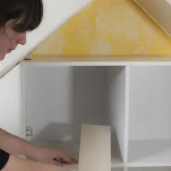 Une femme assemble une maison de poupée. Elle pose une pièce en bois dans une étagère blanche, sous un toit jaune à motifs.