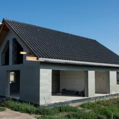 Dom w stanie surowym otwartym, z ciemnym dachem i betonowymi ścianami, czeka na dalsze prace budowlane.
