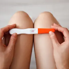 Dłonie trzymające test ciążowy z dwoma kreskami, na tle nóg.