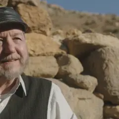 Starszy mężczyzna w czapce i kamizelce, z siwą brodą, uśmiecha się na tle kamiennego muru. Scena z filmu "Uśmiech losu".