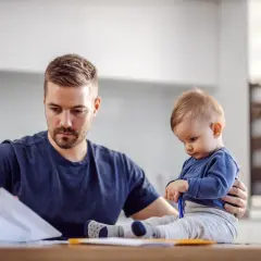 Ojciec pracuje w domu, a dziecko siedzi obok, bawiąc się długopisem. To obraz codziennych kontaktów z dzieckiem w miejscu zamieszkania matki.