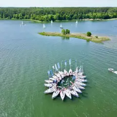 Wielka grupa żaglówek tworzy okrąg na spokojnych, zielonych polskich jeziorach. W tle las i mała wyspa.