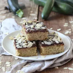 Saftiger Zucchini Kuchen mit Schokoladenglasur und Mandelblättchen, garniert mit frischen Zucchini im Hintergrund.