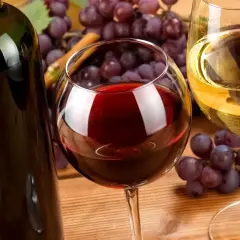 Wybór między wino białe czy czerwone? Kieliszek czerwonego wina obok butelki, a w tle winogrona i kieliszek białego wina.