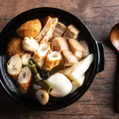 A steaming pot of oden, a Japanese fish cake stew, with various ingredients like daikon and pork belly, served with a wooden spoon.
