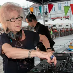Starsza pani dj z pasją miksuje muzykę na placu miejskim, otoczona ludźmi i kolorowymi flagami.