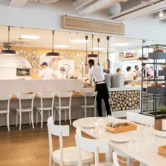 Un restaurant italien moderne avec un four à pizza visible. Des chefs préparent des plats derrière le comptoir, tandis que des tables sont dressées pour les clients.