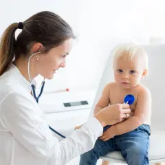 Lekarka bada małego chłopca stetoskopem. Dziecko siedzi na krześle, ubrany w dżinsy. Badanie jest rutynowe, nie ma mowy o wnętrostwie.