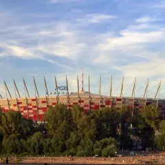 Stadion Narodowy w Warszawie, z imponującą pojemnością, otoczony zielenią, na tle błękitnego nieba z chmurami. Ludzie odpoczywają na plaży.