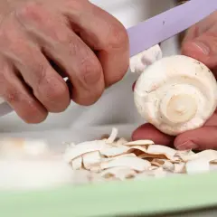 Dłonie obierają pieczarkę nożem. Czy pieczarki trzeba obierać? Skórka jest usuwana, tworząc stos cienkich plasterków.