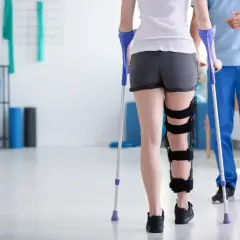 Pacjentka z ortezą na nodze uczy się chodzić o kulach pod okiem fizjoterapeuty w gabinecie rehabilitacyjnym.