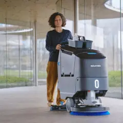 Mujer operando una máquina de Nilfisk, parte de sus avanzados sistemas de limpieza, en un pasillo moderno con grandes ventanales.