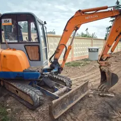 Pomarańczowa koparka Hitachi z gąsienicami i szerokim lemieszem do koparki, gotowa do pracy na budowie.