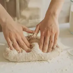 Manos amasando masa pan sobre una mesa con harina esparcida. Un frasco de vidrio con harina y un cuenco están al fondo.
