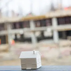 Biały model domu stoi na pierwszym planie, w tle widać budowę domu wielorodzinnego.