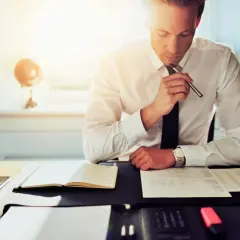 Mężczyzna w białej koszuli i krawacie siedzi przy biurku, trzymając długopis i analizując dokumenty.