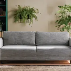 Sofa w kolorze popielaty jaki to kolor, otoczona zieloną biblioteczką i bujną roślinnością.