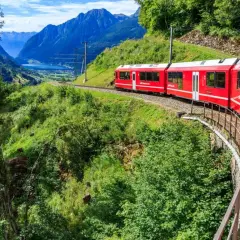 Röda tåget Bernina Express slingrar sig genom ett grönt alplandskap med en sjö och berg i bakgrunden. Undrar du över bernina express pris?